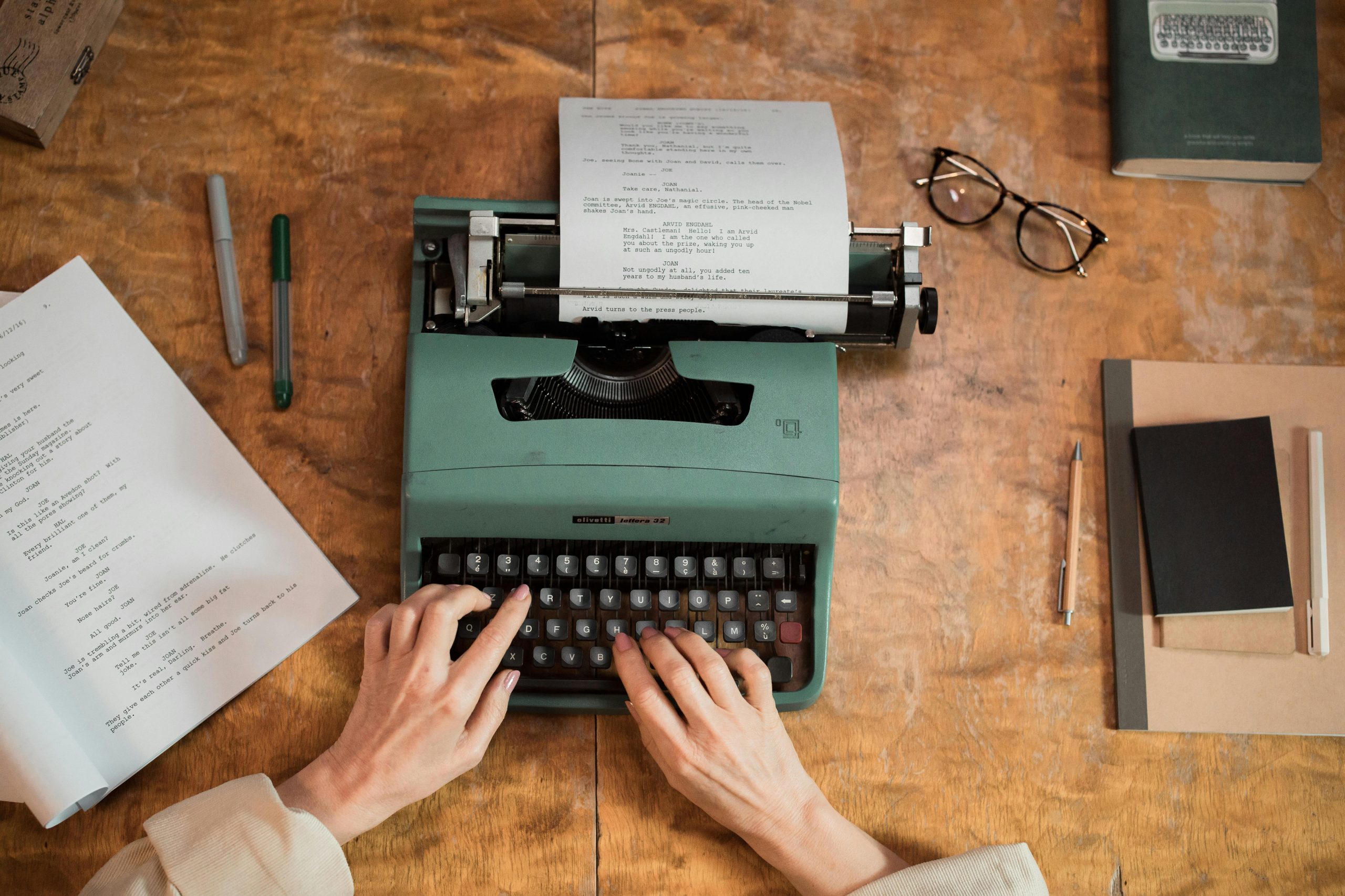 a set of hands working at a green typewriter, while paper, pens, glasses and cups of coffee litter the wooden desktop